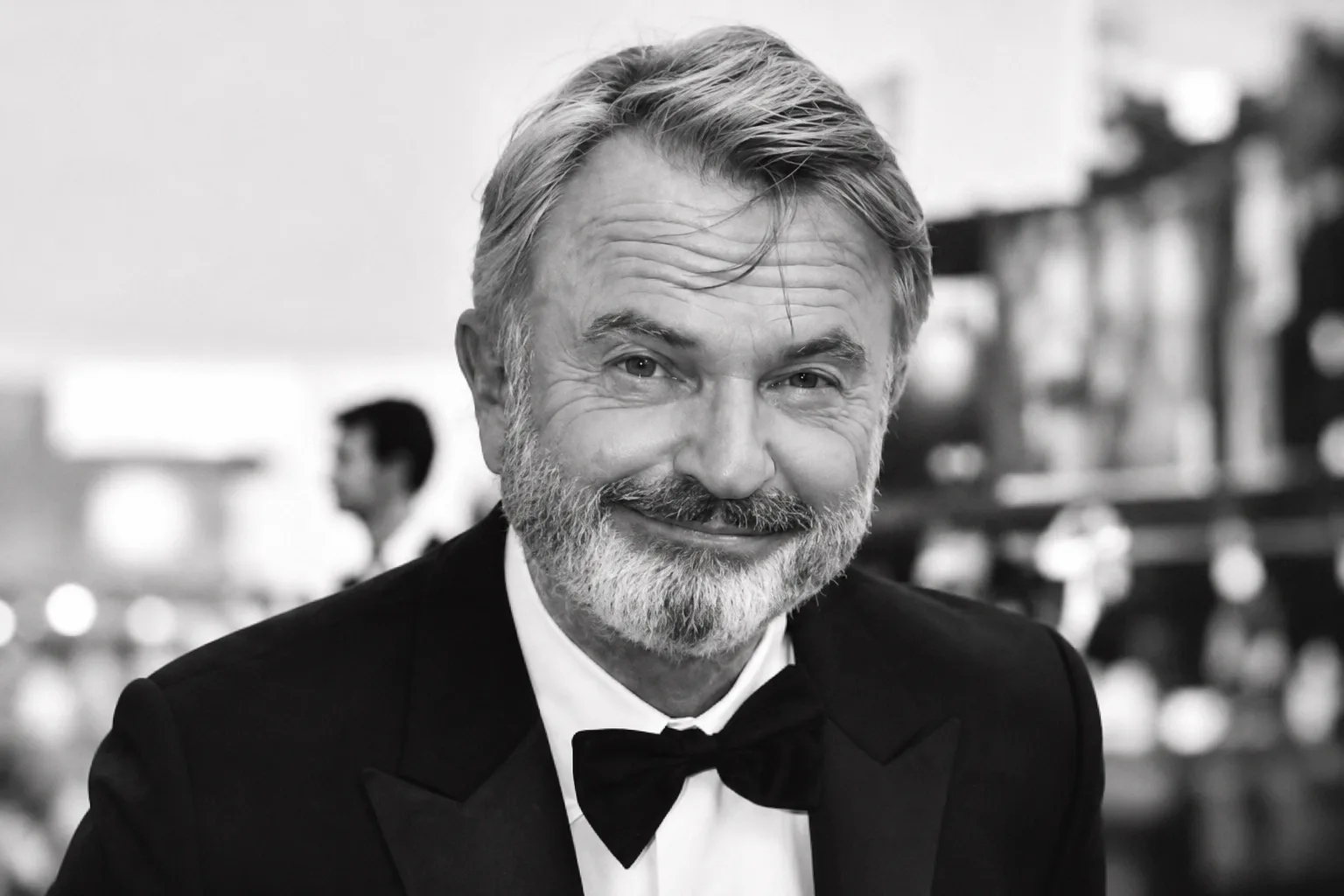 Black-and-white portrait of Sam Neill smiling in a tuxedo, used for coverage of the acclaimed actor's death at age 78.