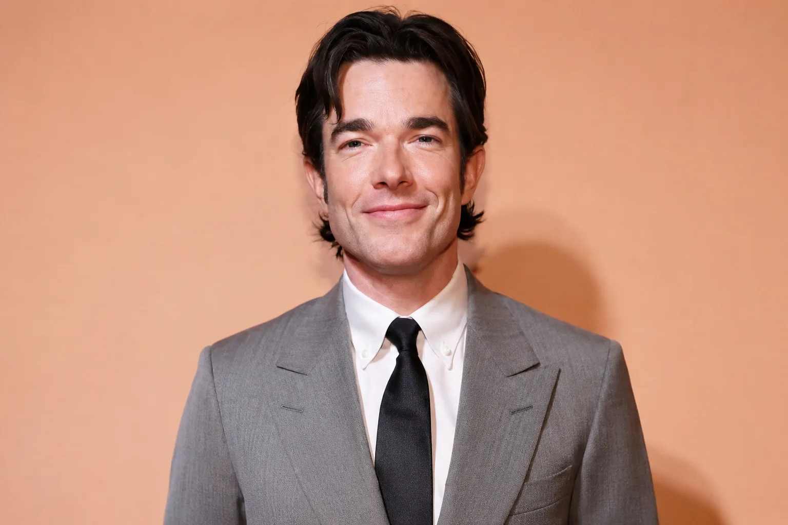 John Mulaney wearing a light gray suit and black tie while posing against a warm peach-colored backdrop in a professional event portrait.