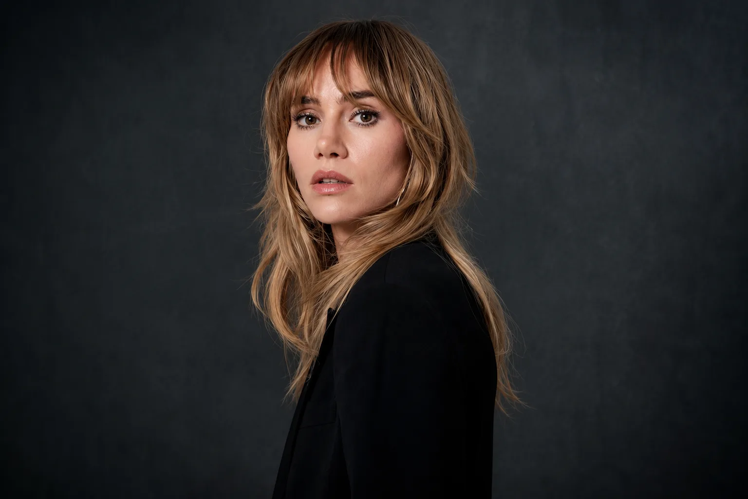 Suki Waterhouse poses in a black blazer against a dark studio backdrop during a promotional portrait for her album Loveland.