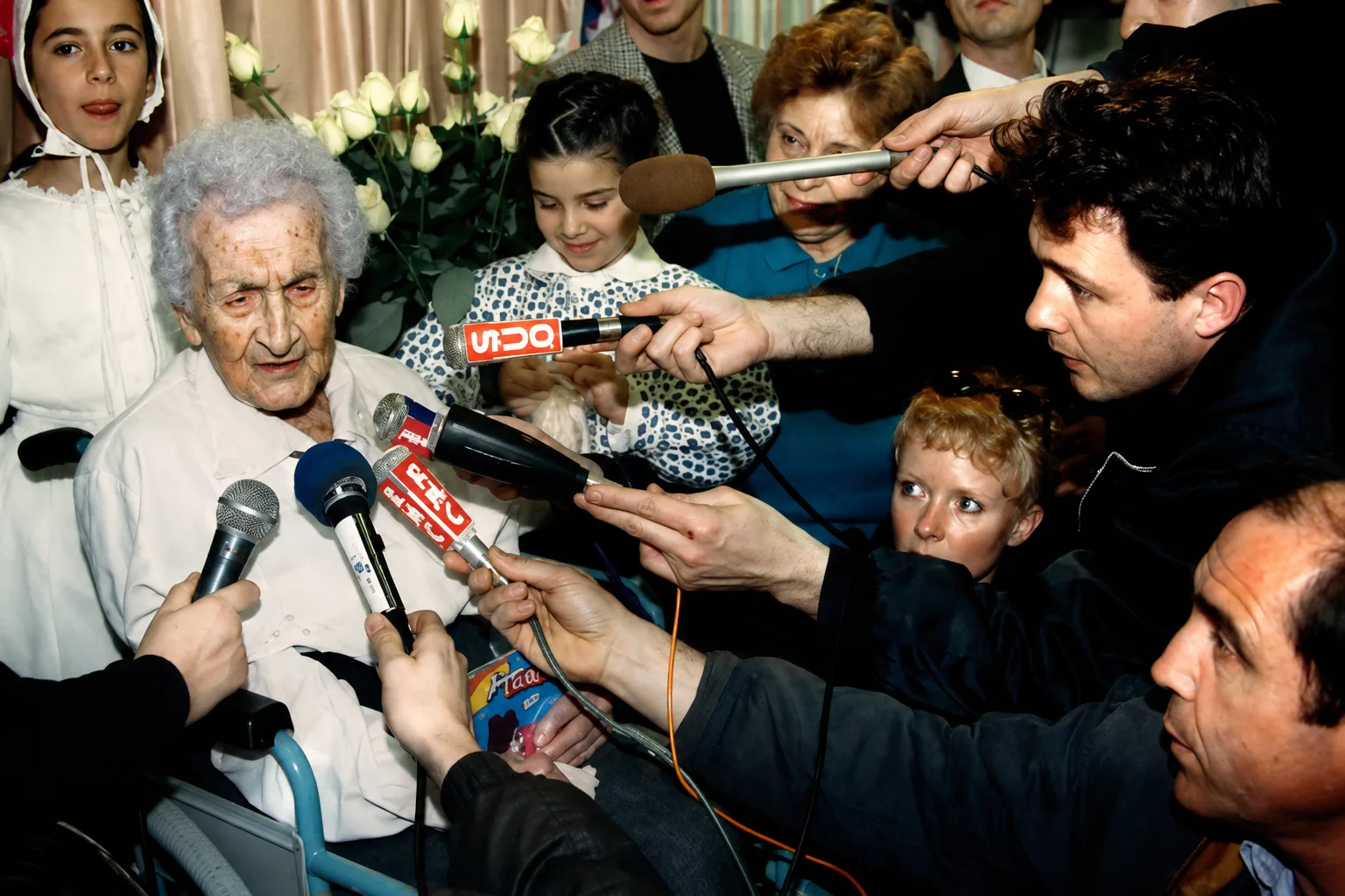 An elderly supercentenarian speaks to reporters during a press event, reflecting the themes of Sam Green's documentary The Oldest Person in the World.